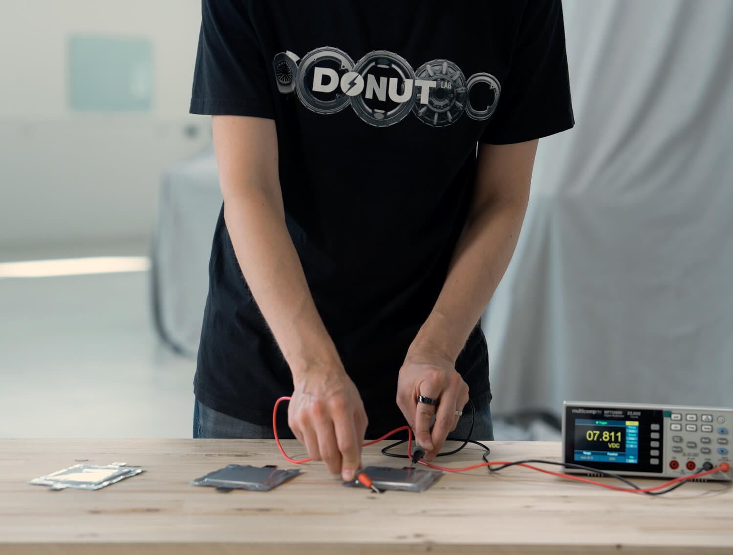 A person in a "Donut" shirt uses probes to test metal plates connected to an electronic meter on a wooden table.